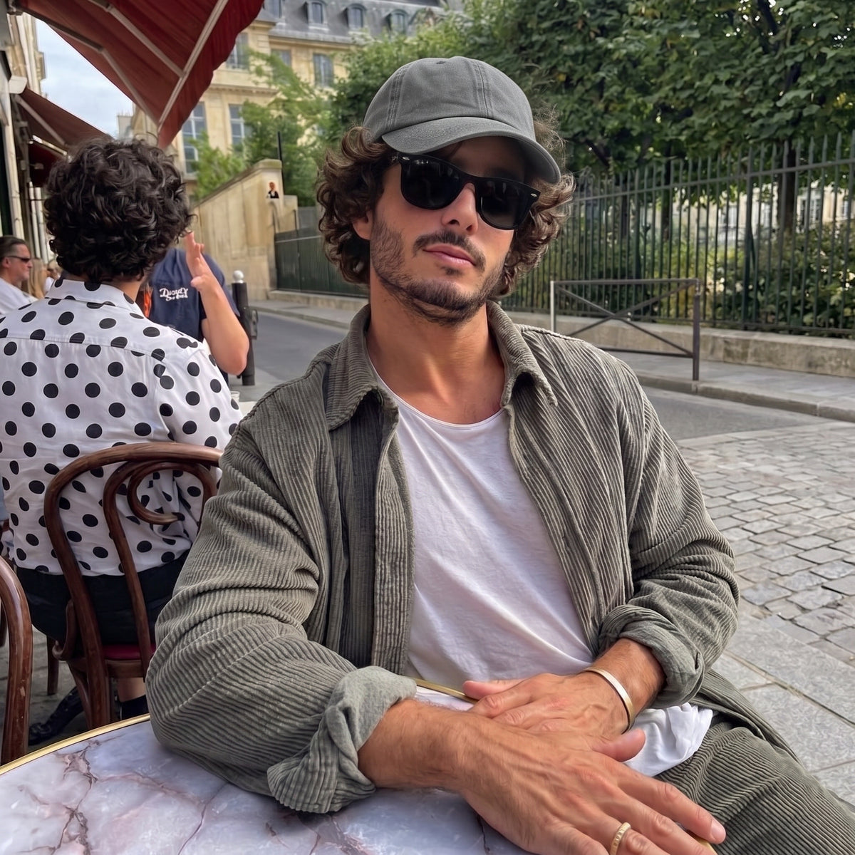 A man sitting at an outdoor cafe table wearing a gray corduroy overshirt, matching gray trousers, a gray baseball cap, and black sunglasses over a white t-shirt.