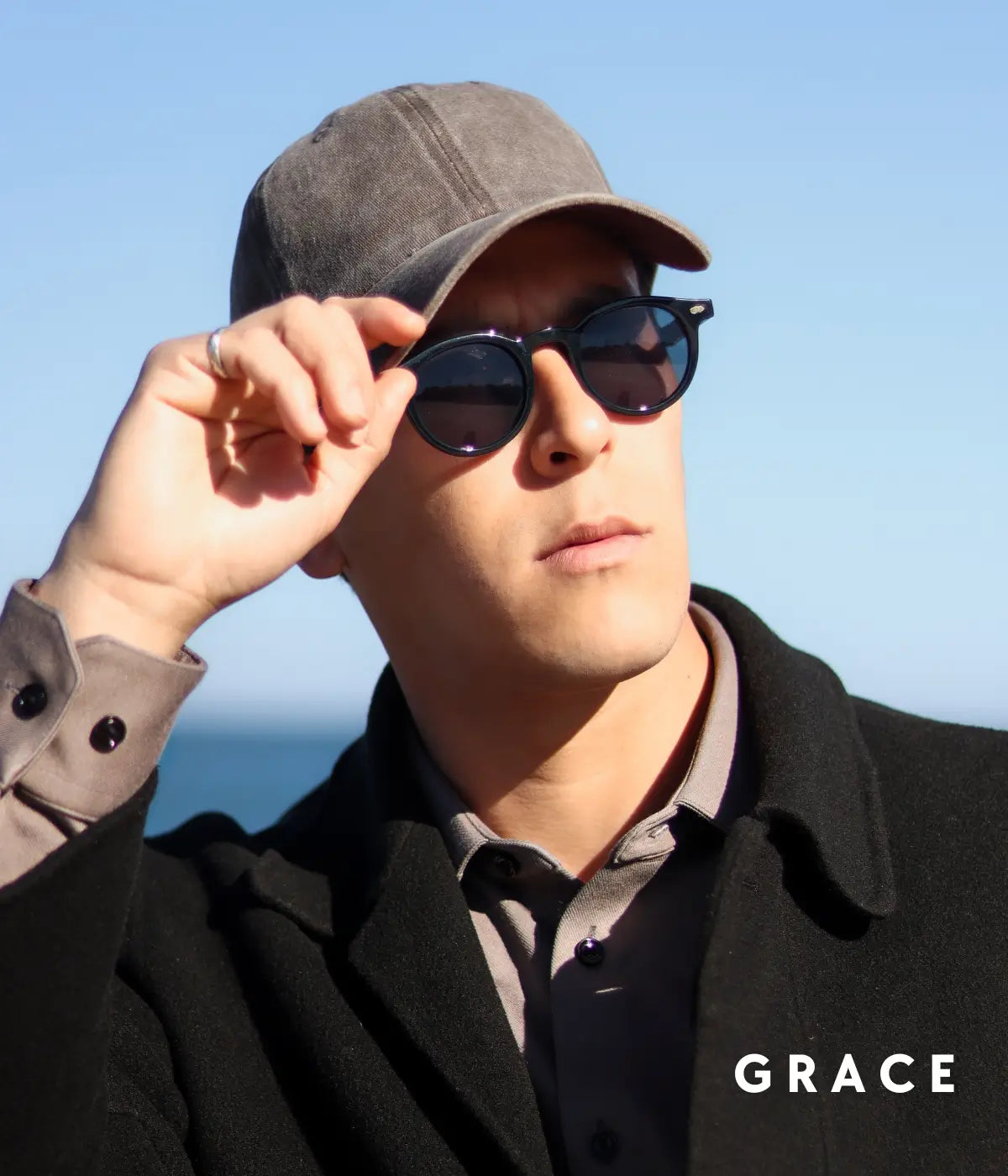 A man outdoors wearing a gray cap, dark round sunglasses, a black coat, and a gray collared shirt, with the brand name GRACE in the bottom right corner.