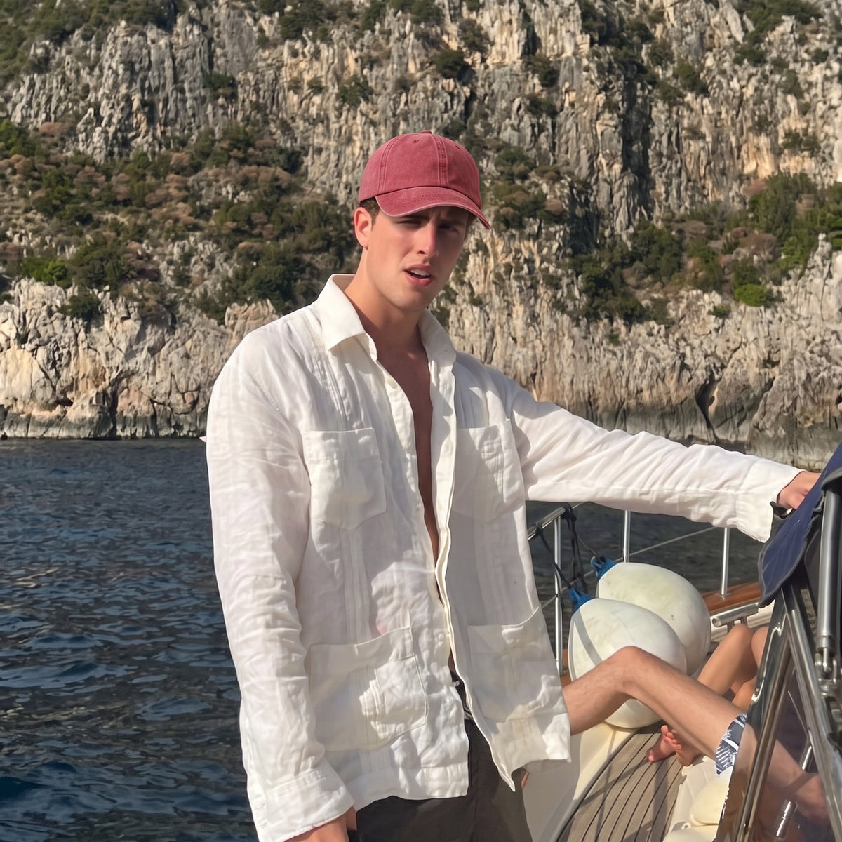 A man wearing a white linen shirt and a pink cap stands on a boat with a rocky coastline in the background.