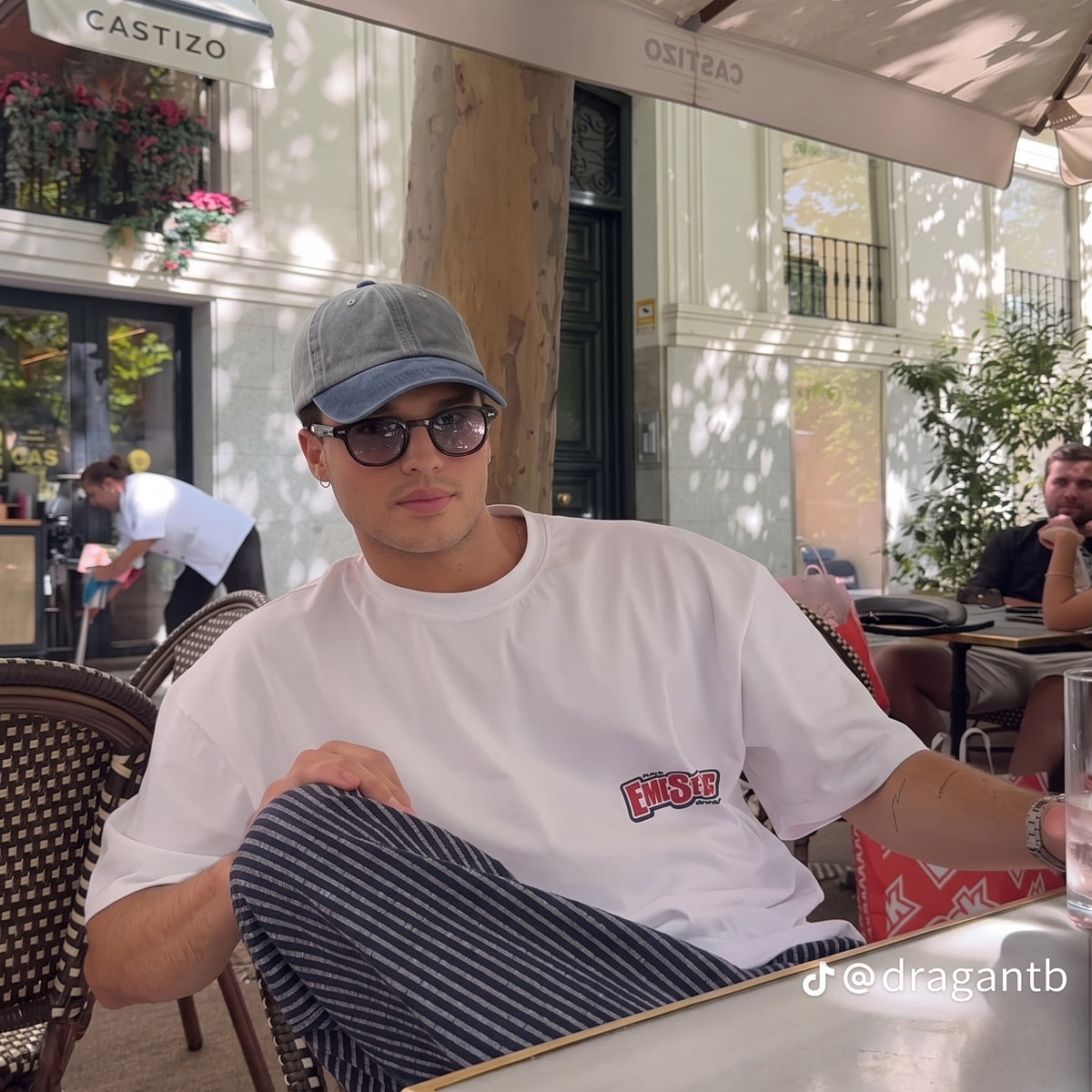 A man sitting at an outdoor cafe wearing a white t-shirt with a red and blue 'Nube' logo on the chest, a gray baseball cap, and striped pants.