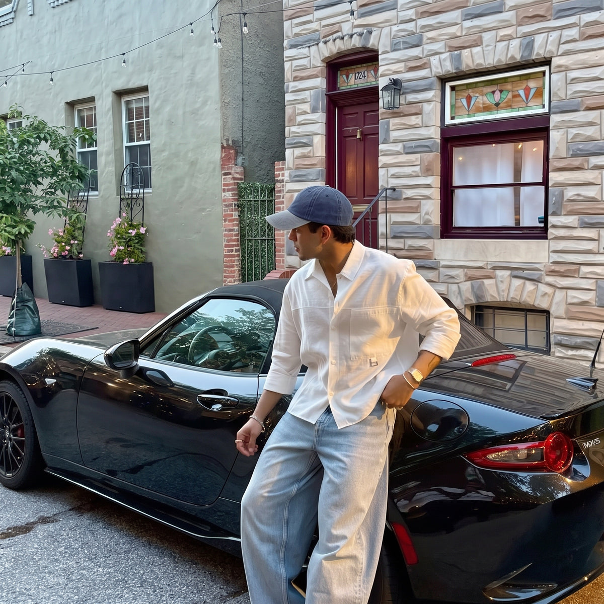 A man wearing a white button-down shirt, light blue jeans, and a blue baseball cap leans against a black convertible sports car parked in front of a stone building.
