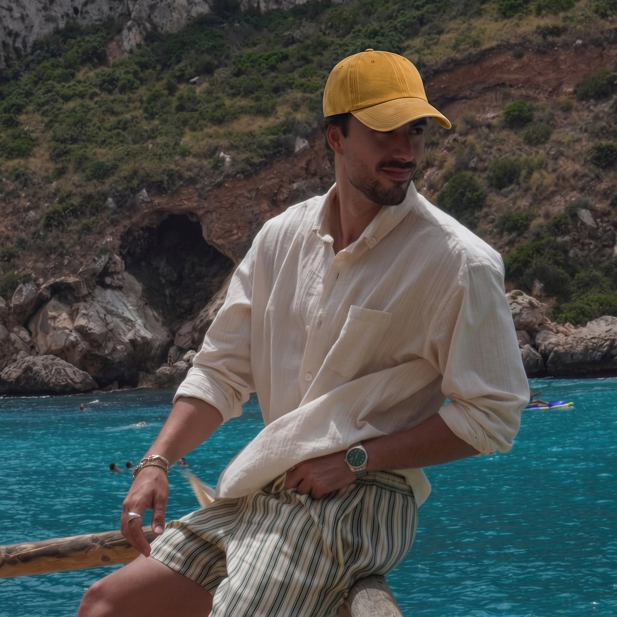 A man wearing a yellow baseball cap, a cream linen button-up shirt, and striped shorts sits on a wooden railing overlooking a scenic coastal landscape with turquoise water and rocky cliffs.