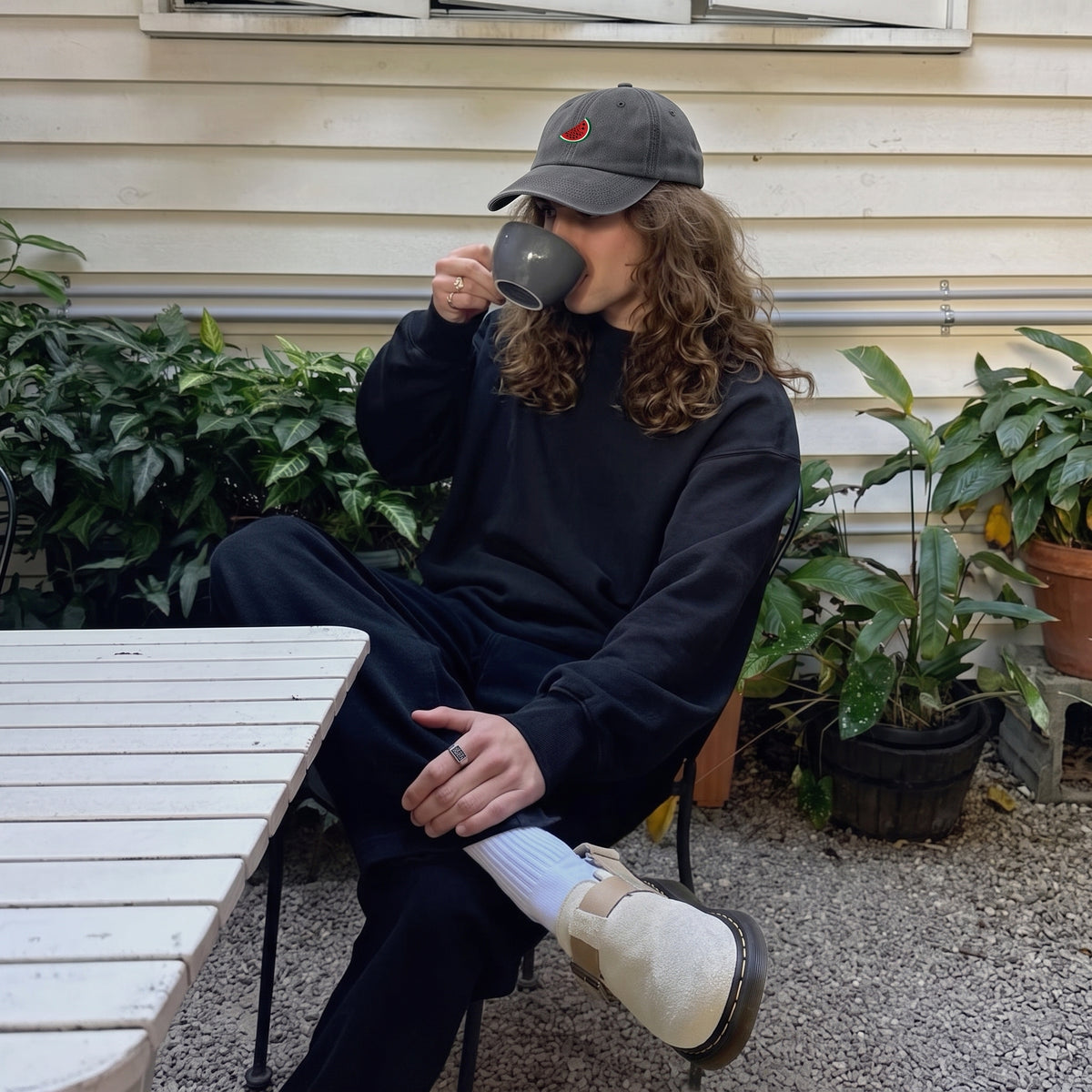 A person with curly hair sits on a chair outdoors, wearing a dark navy sweatshirt and matching pants, a dark gray baseball cap, white socks, and a beige shoe, while holding a dark gray mug. The background shows a beige house exterior with plants and a white table.