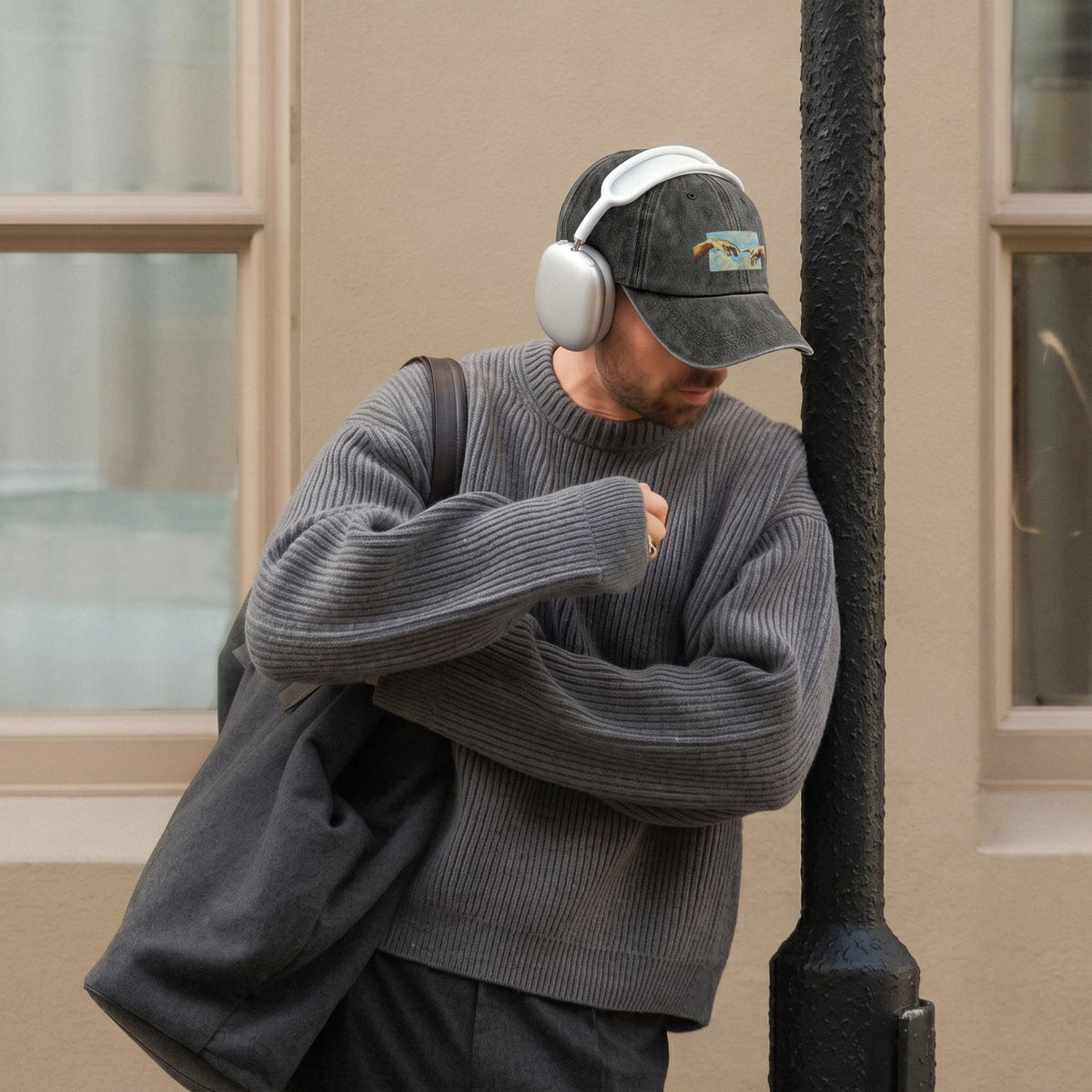 A man wearing a dark gray ribbed knit sweater, a black cap with a colorful graphic, and white over-ear headphones leans against a black pole next to a beige building wall.