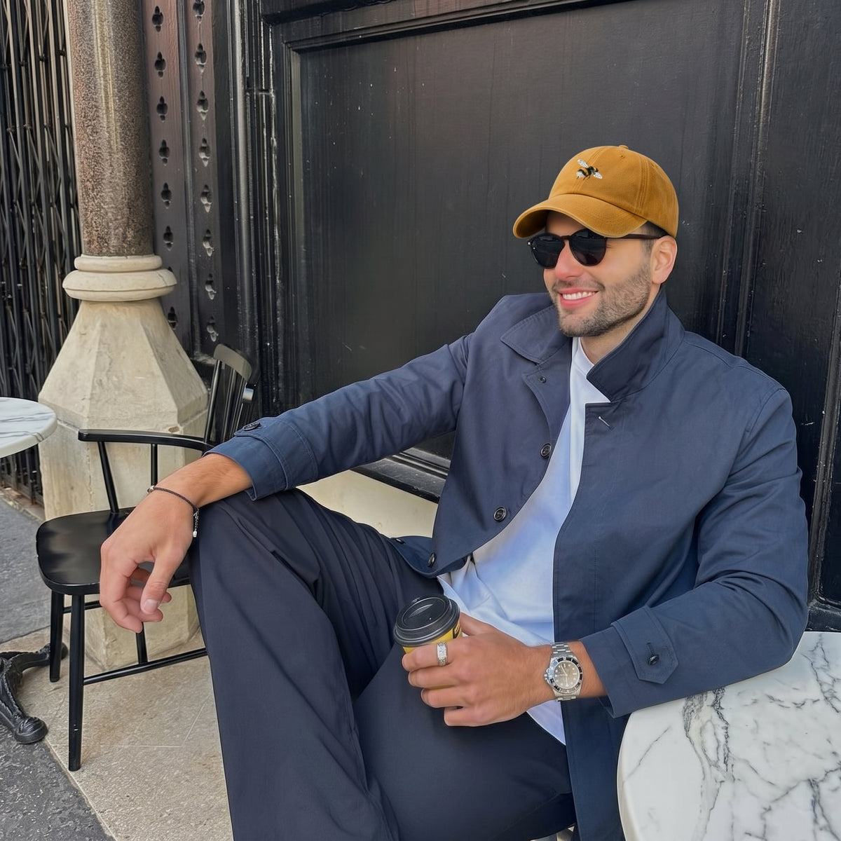 A man wearing a navy blue jacket and trousers, a light blue shirt, a mustard yellow baseball cap, and sunglasses sits outdoors on a cafe patio holding a coffee cup.
