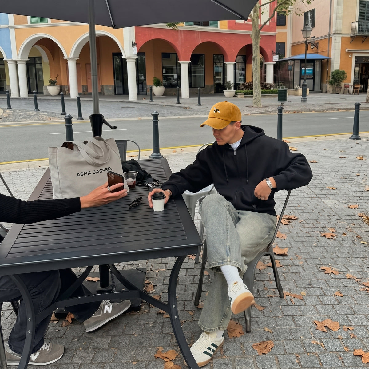 A man in a black hoodie, light wash jeans, white socks, and a yellow cap sits at an outdoor cafe table holding a coffee cup, with another person holding a phone visible on the left. A beige tote bag with 'ASHA JASPER' printed on it rests on the table.