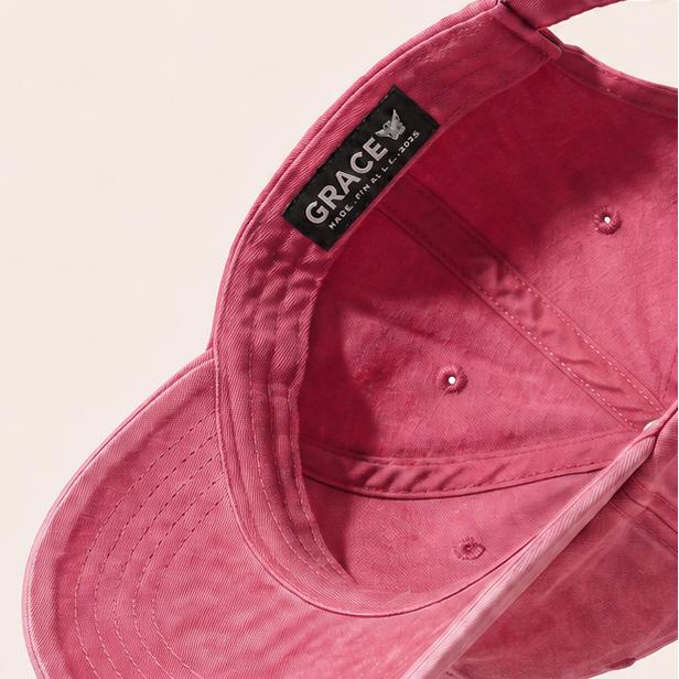 A dusty rose pink cotton baseball cap shown floating at an angle against a warm cream background, with a black interior label reading 'GRACE'.
