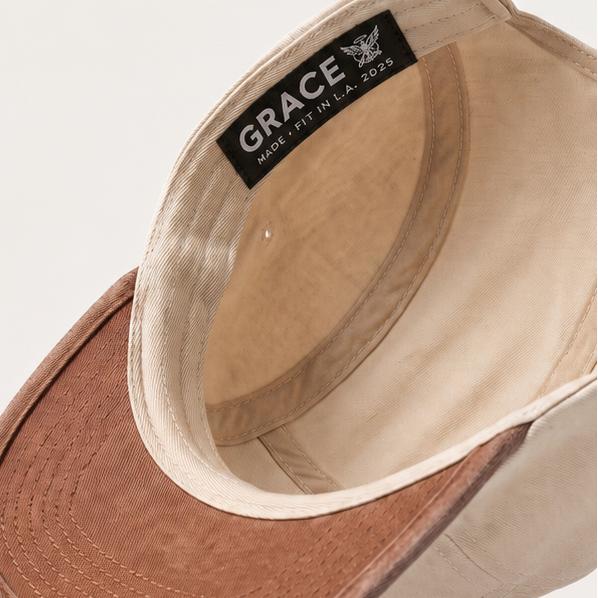 A beige baseball cap with a brown suede-look visor, shown floating at an angle against a light beige background. The inside of the crown features a black brand label reading 'GRACE'.