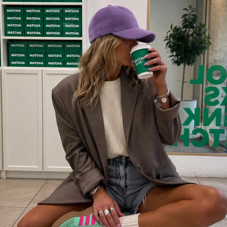 A woman sits cross-legged on a marble bench in a cafe, wearing a purple baseball cap, a brown blazer, a cream t-shirt, denim shorts, and mint green sneakers. She is holding a green coffee cup.