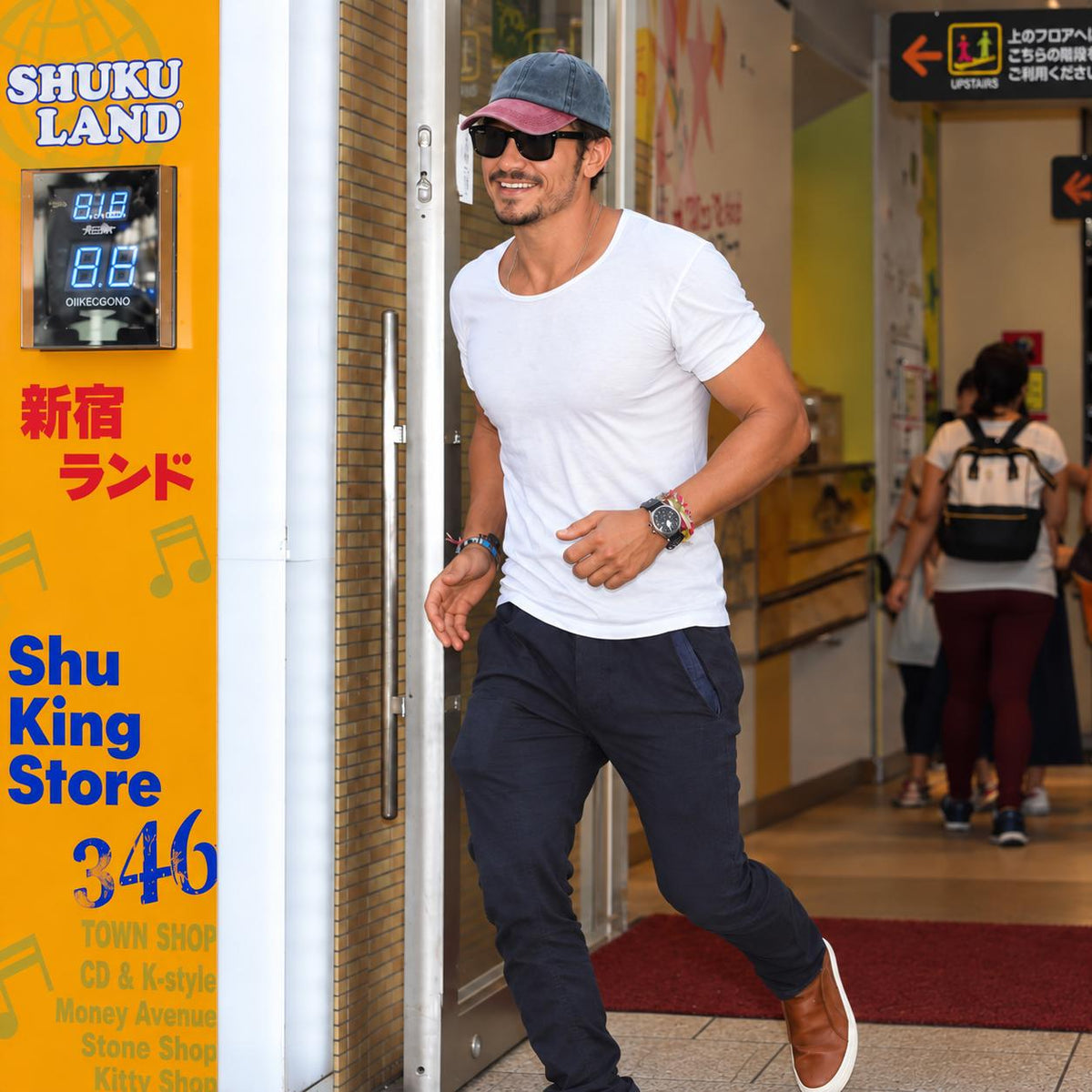 A man wearing a white t-shirt, dark navy pants, a blue cap, and sunglasses walks out of a Shuku Land store. The storefront features a bright yellow sign with Japanese text and a digital clock.