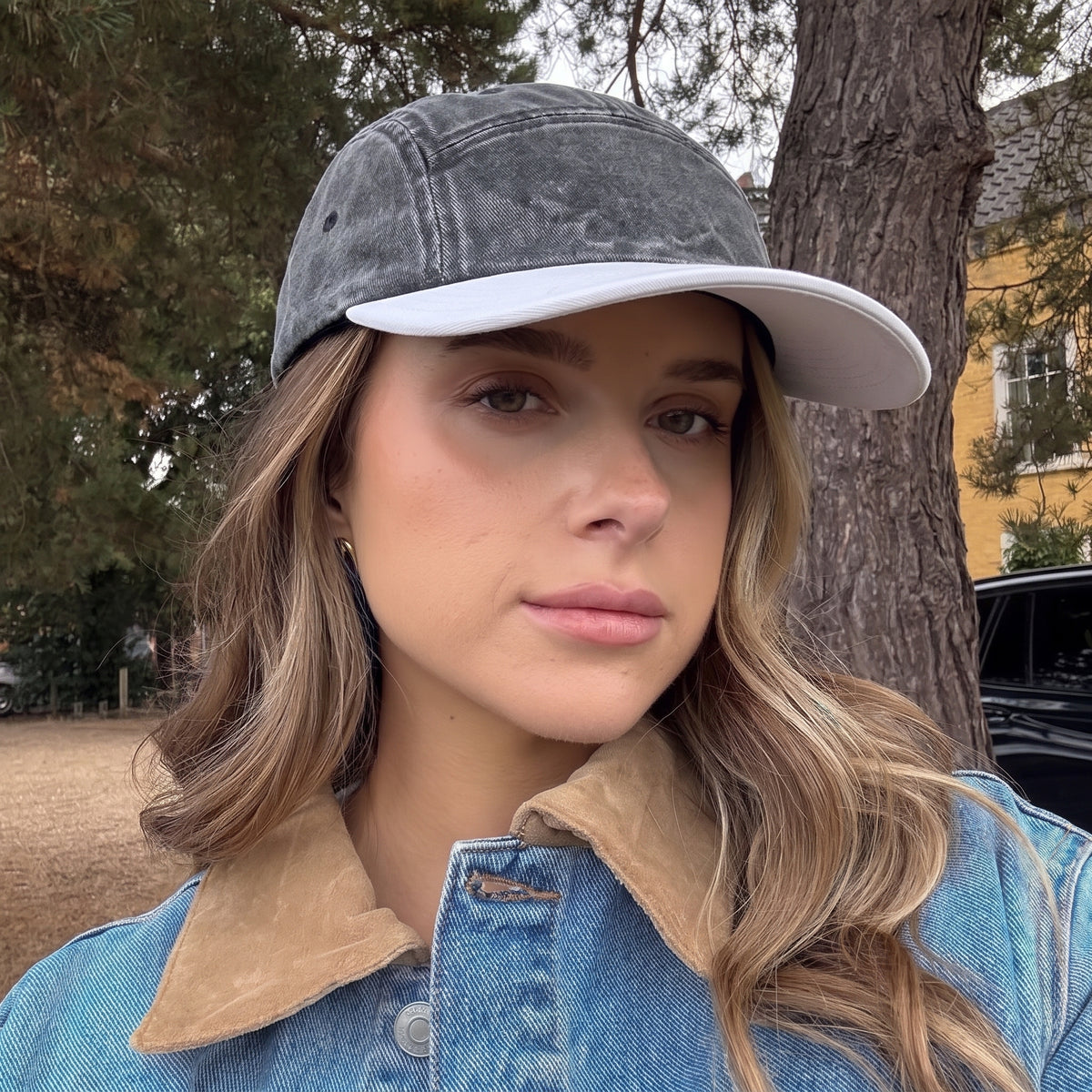 A woman wearing a washed dark gray and white baseball cap, standing outdoors in front of a tree and a yellow building.
