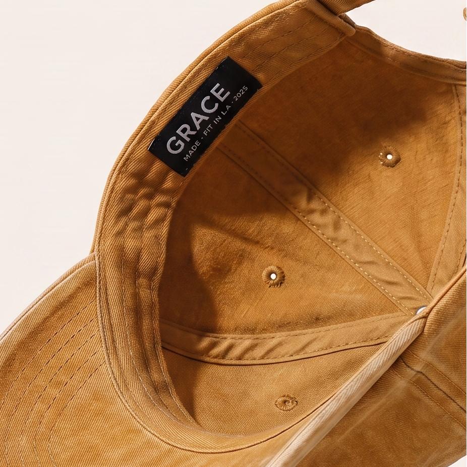 A tan cotton cap with a curved brim, shown floating at an angle against a light beige background. The inside of the crown features a black label with the brand name 'GRACE' in white text.