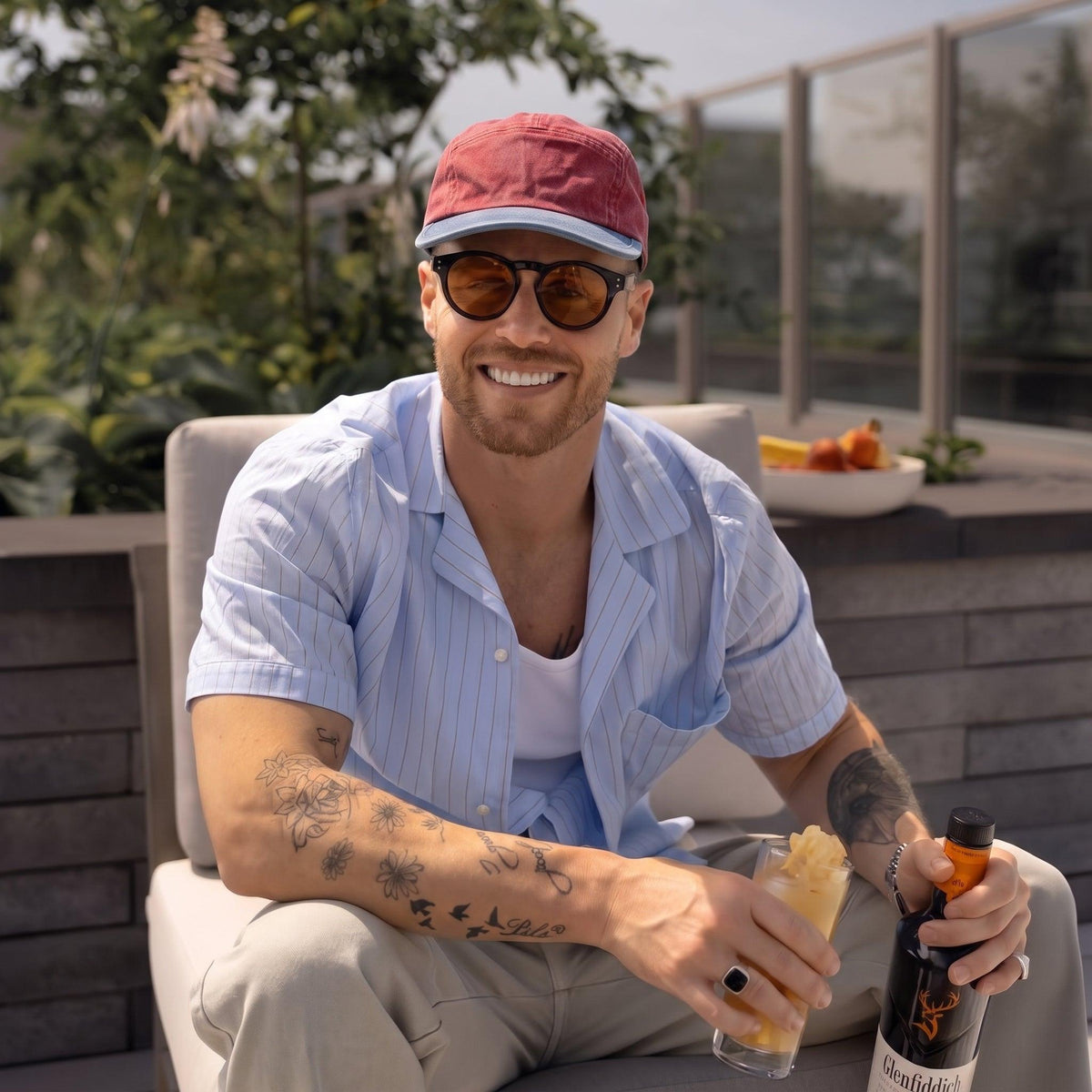 A smiling man sitting outdoors on a patio chair, wearing a red cap, round sunglasses, a light blue pinstriped short-sleeve button-up shirt, and beige pants. He is holding a drink and a bottle labeled 'Saffron'.