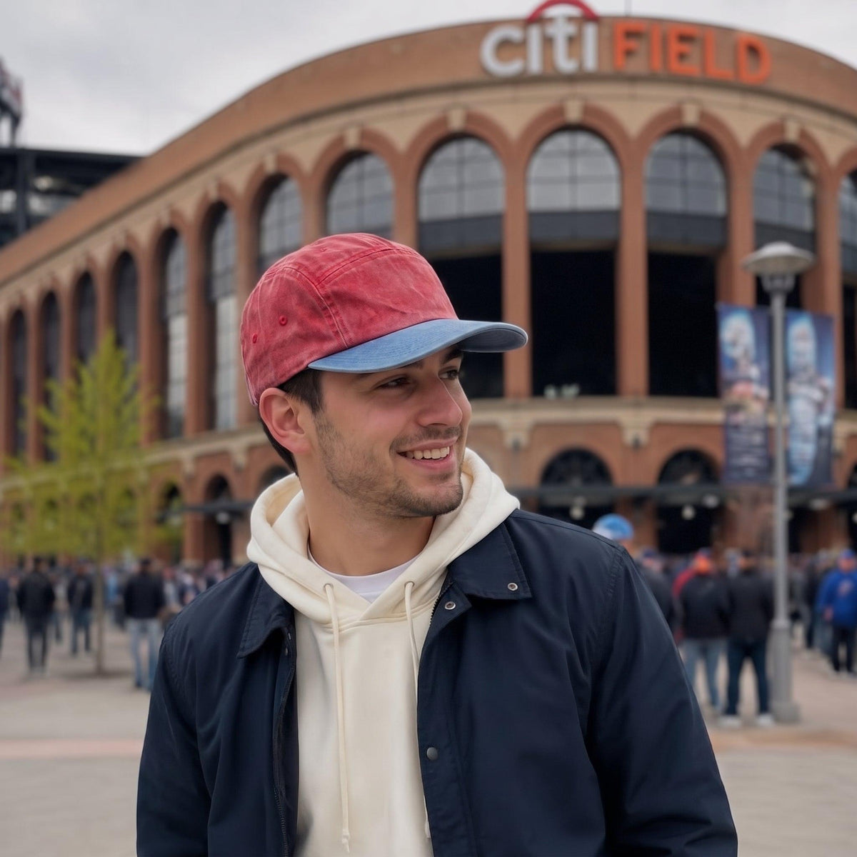 A man wearing a red and blue baseball cap, a cream hoodie, and a dark navy jacket stands in front of Citi Field stadium, smiling and looking to the side.