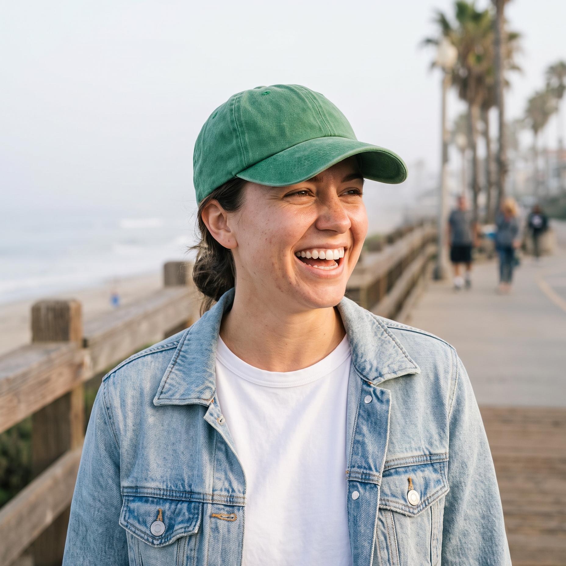 A woman smiling on a wooden boardwalk by the beach, wearing a green baseball cap, a light blue denim jacket, and a white t-shirt. Palm trees and ocean waves are visible in the background.