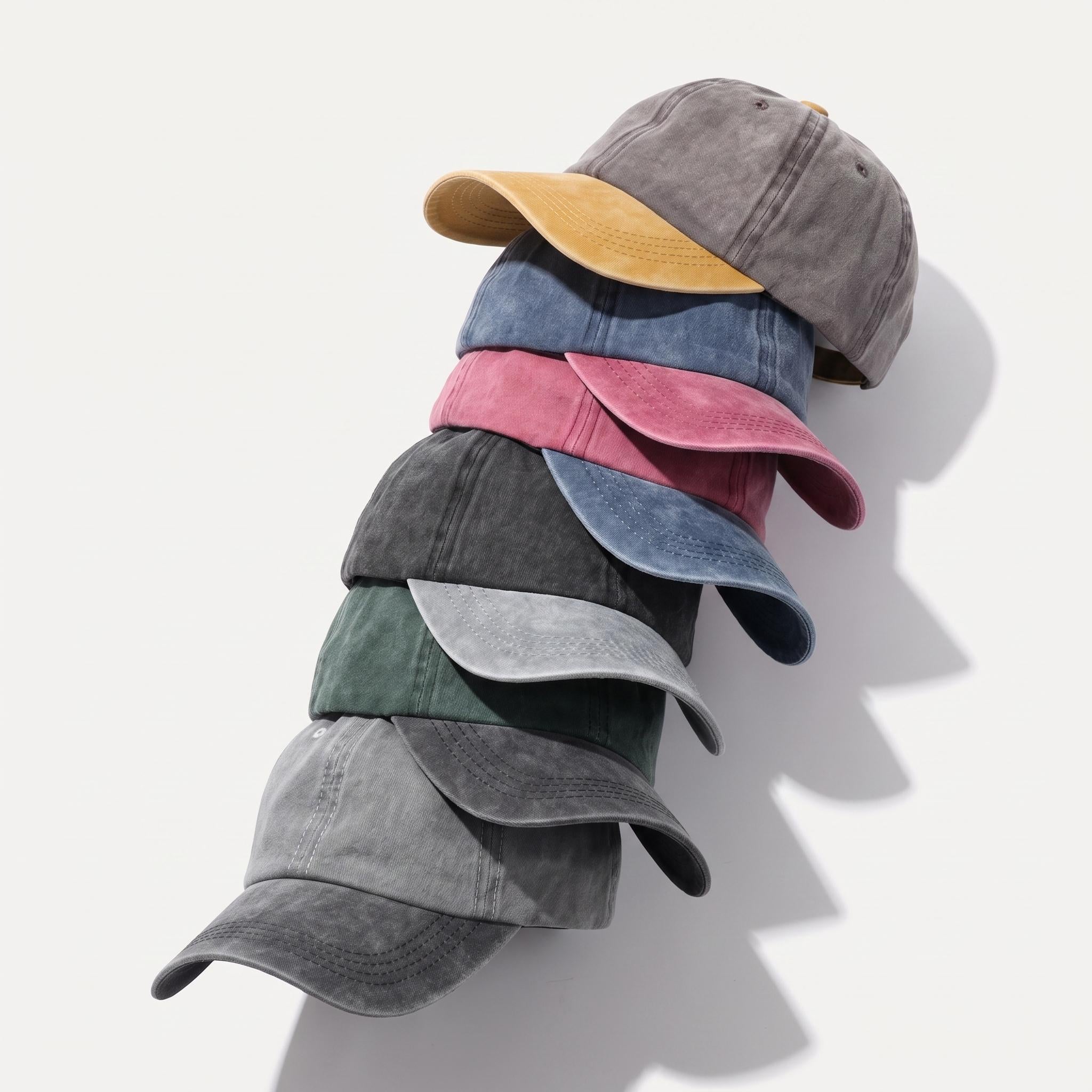 A stack of six baseball caps in different muted colors, including gray, mustard yellow, blue, pink, charcoal, and sage green, arranged diagonally on a light off-white background with soft shadows.