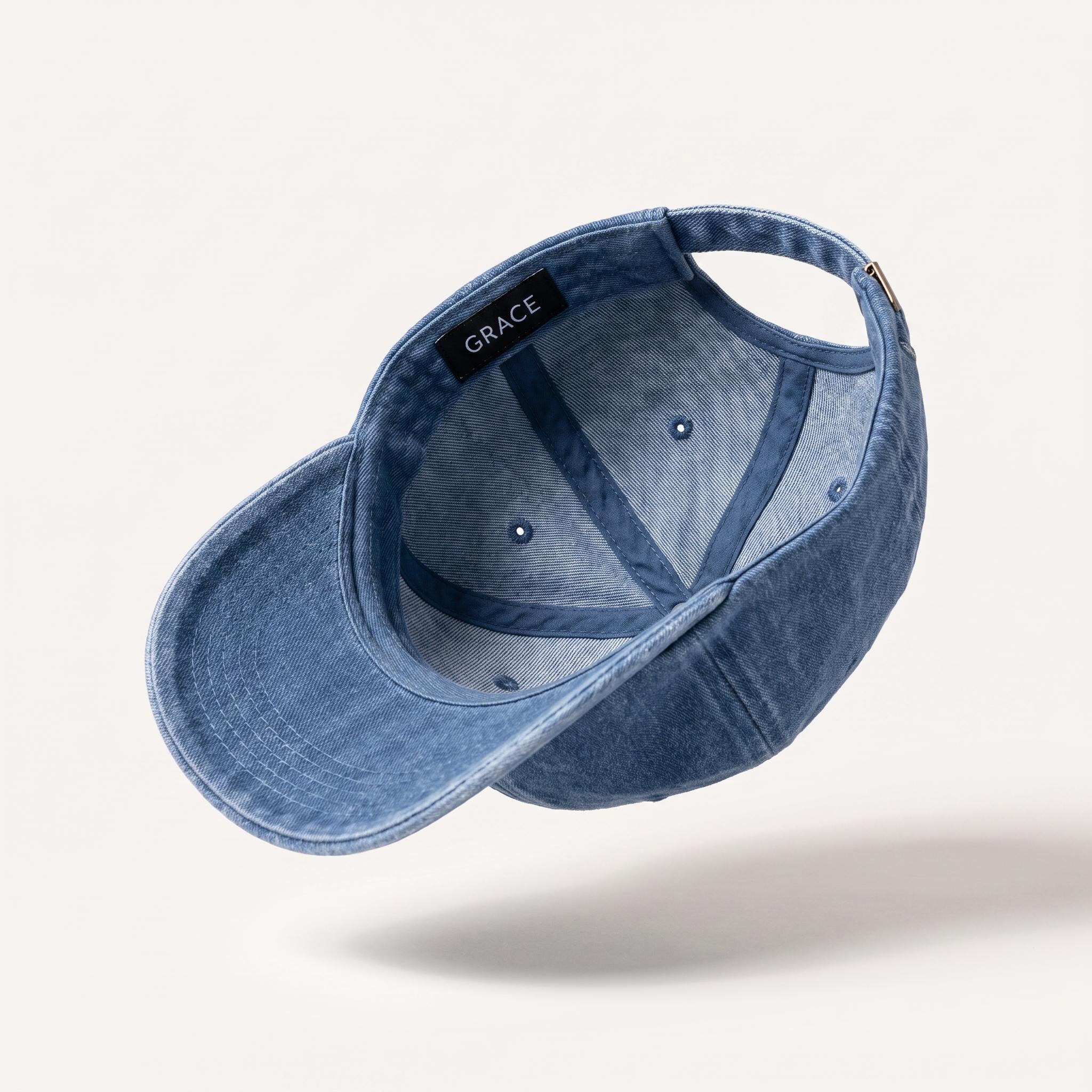 A blue denim baseball cap floating at an angle against a light beige background, showing the interior with a black brand label that reads 'GRACE'.