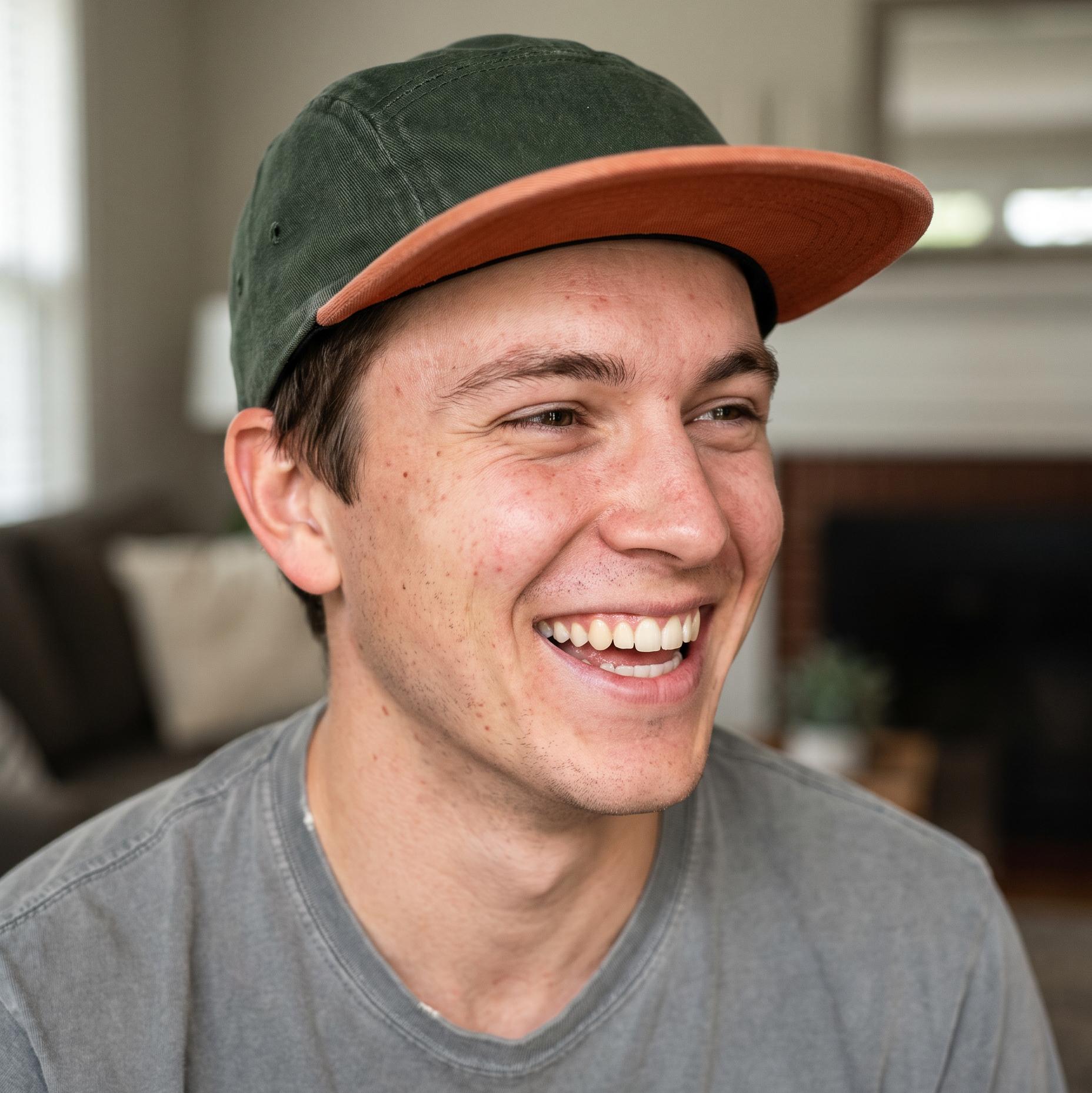 A smiling young man wearing a green baseball cap with a brown visor and a gray t-shirt, photographed in a brightly lit indoor setting.