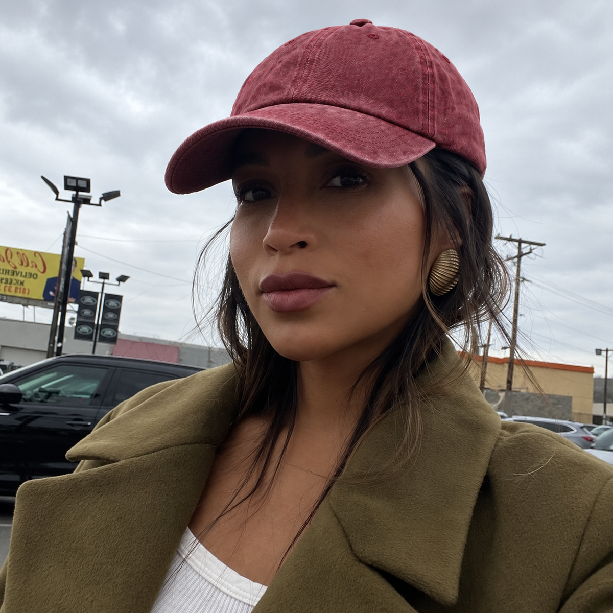 A woman wearing a dusty rose pink corduroy baseball cap and an olive green coat, photographed outdoors in a cloudy sky with a parking lot and store in the background.