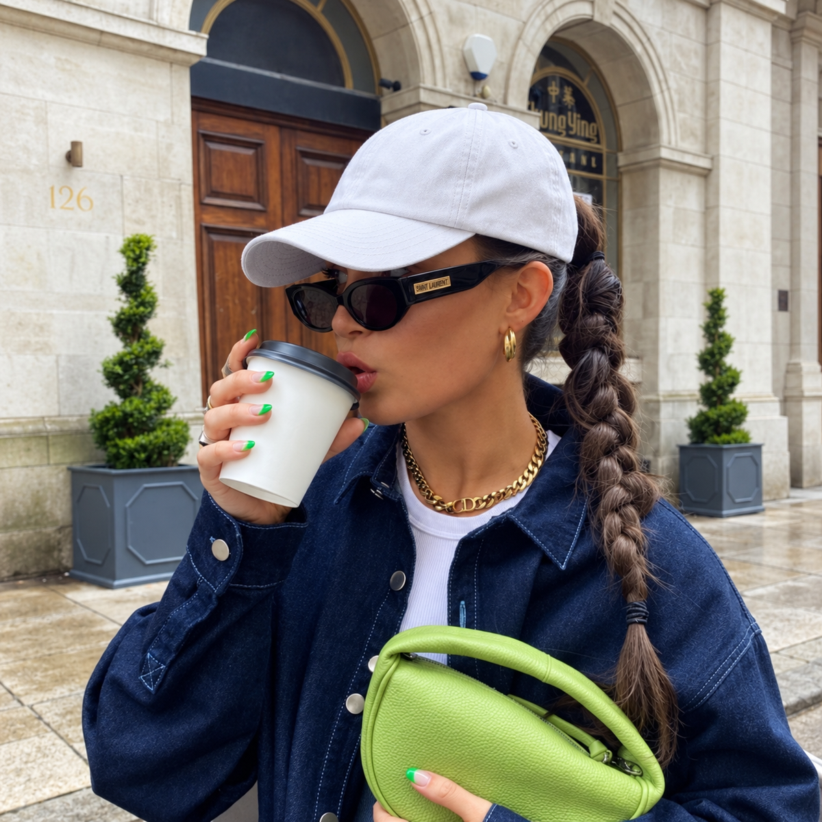 A woman wearing a white baseball cap, black sunglasses, a blue denim jacket, and a white t-shirt holds a white coffee cup and a lime green pouch bag, standing in front of a stone building.