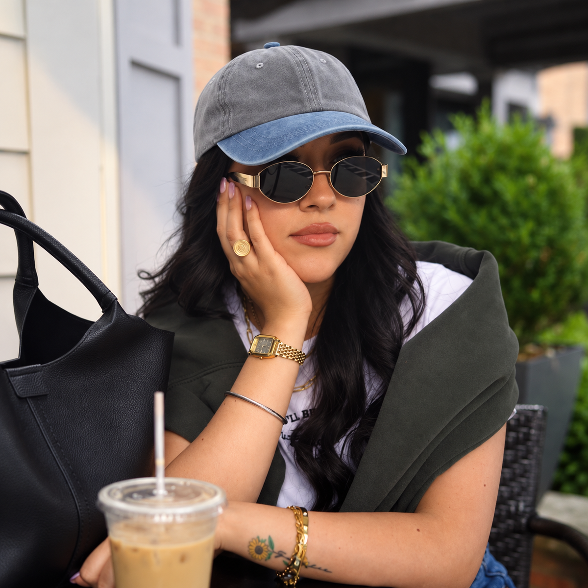 A woman sitting at an outdoor cafe table wearing a gray and blue baseball cap, round sunglasses, a white t-shirt, a dark green cardigan, and multiple gold-toned bracelets and a watch. She has a black tote bag and an iced coffee in the foreground.
