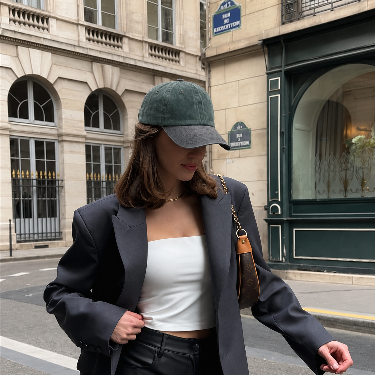 A woman wearing a dark green baseball cap, a black blazer, a white tube top, and black pants, carrying a brown shoulder bag, standing on a city street.