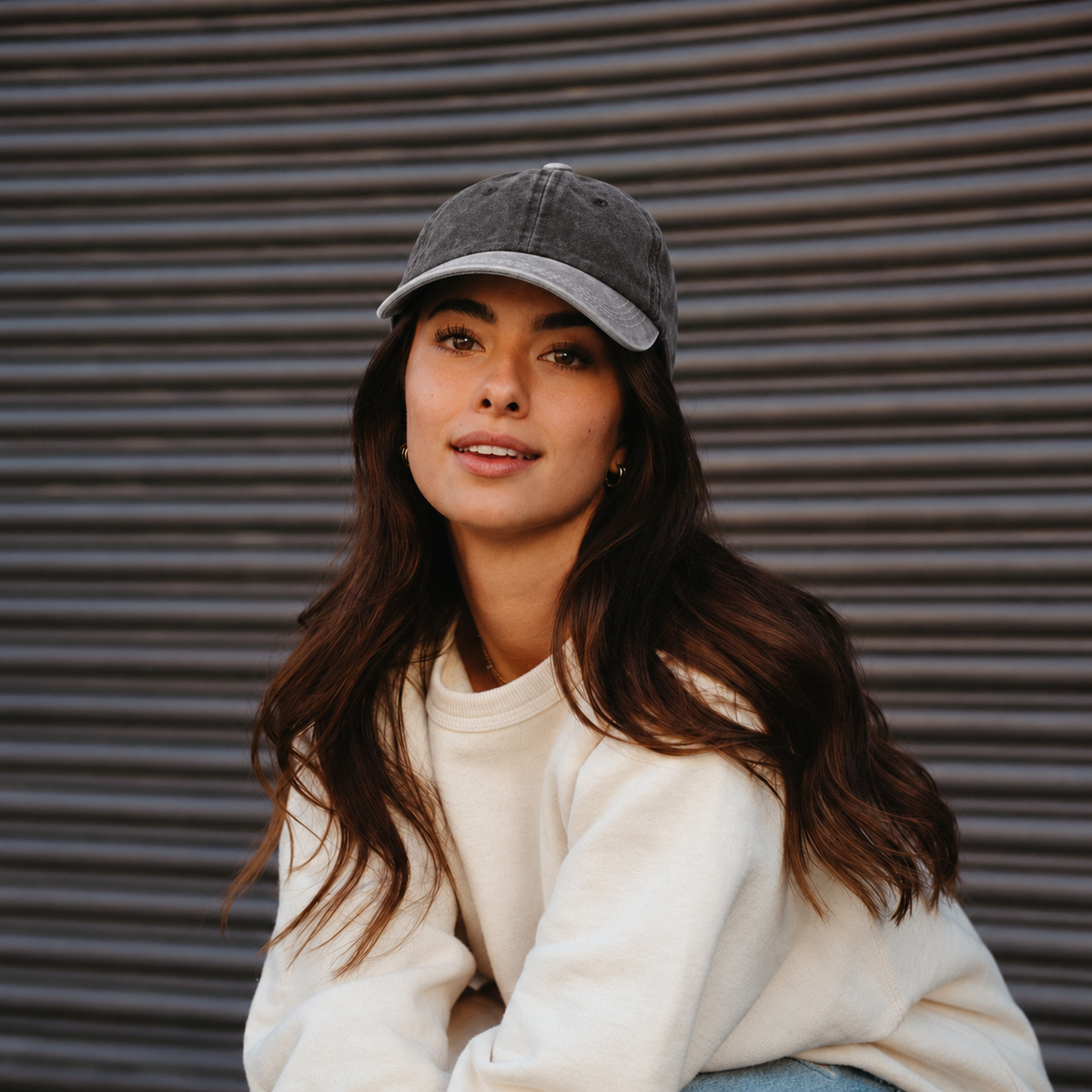 A woman wearing a gray baseball cap and a cream-colored sweatshirt, posing in front of a dark gray metal shutter.