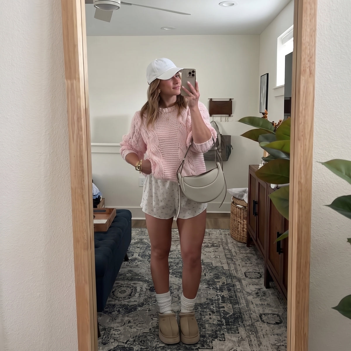 A woman takes a mirror selfie in a home setting, wearing a light pink knit sweater, light gray patterned shorts, a white baseball cap, white socks, tan slip-on shoes, and a light beige crescent shoulder bag.