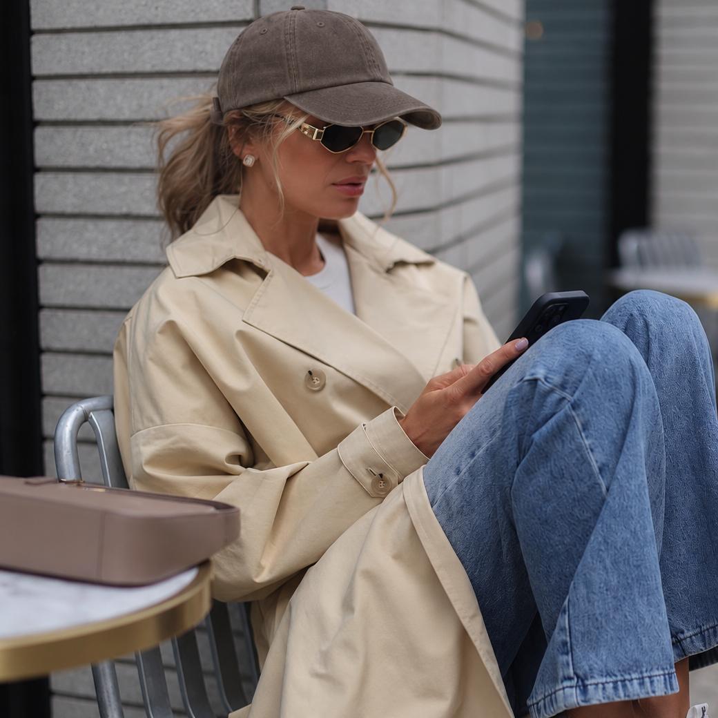 A woman sits on a chair outdoors, wearing a beige trench coat, blue jeans, a brown baseball cap, and sunglasses while looking at her phone. A matching beige box rests on a table beside her.