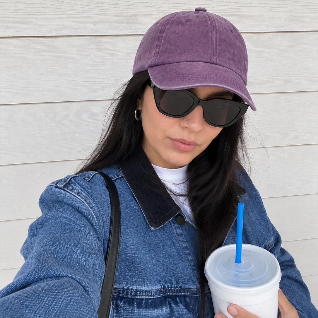 A woman wearing a purple baseball cap and black sunglasses, posing in a blue denim jacket and holding a white cup with a blue straw against a white wood-paneled wall.