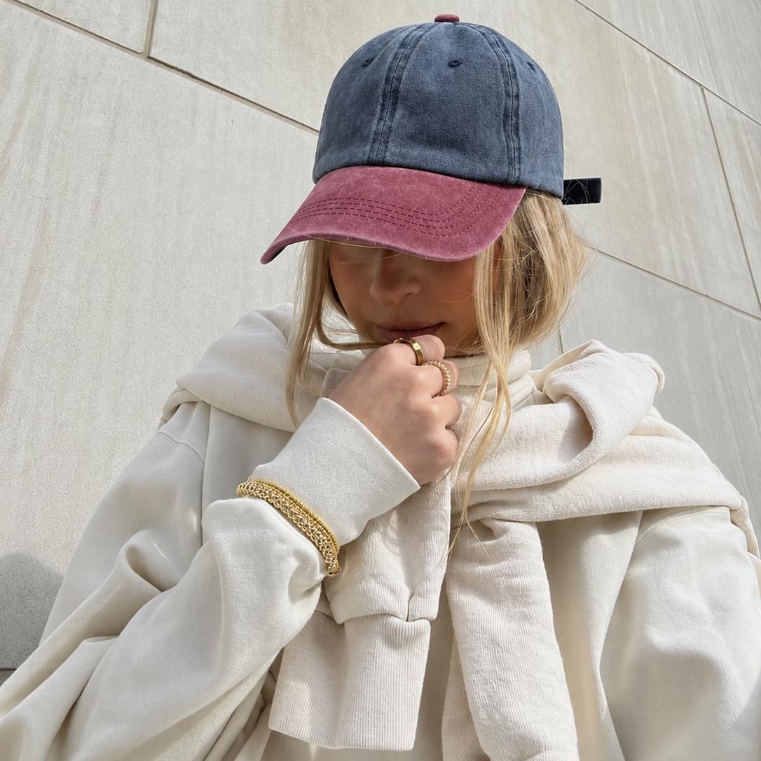 A person wearing a washed blue and dusty rose baseball cap, a cream-colored oversized hoodie, and a gold chain bracelet, standing against a light beige stone wall.