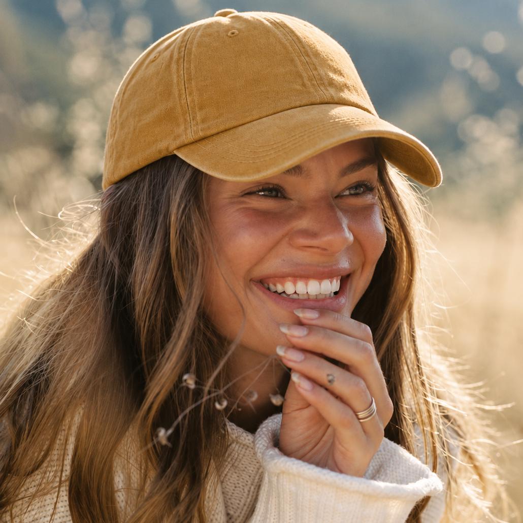 A woman smiling outdoors wearing a mustard yellow baseball cap and a cream knit sweater, with a softly blurred natural background.