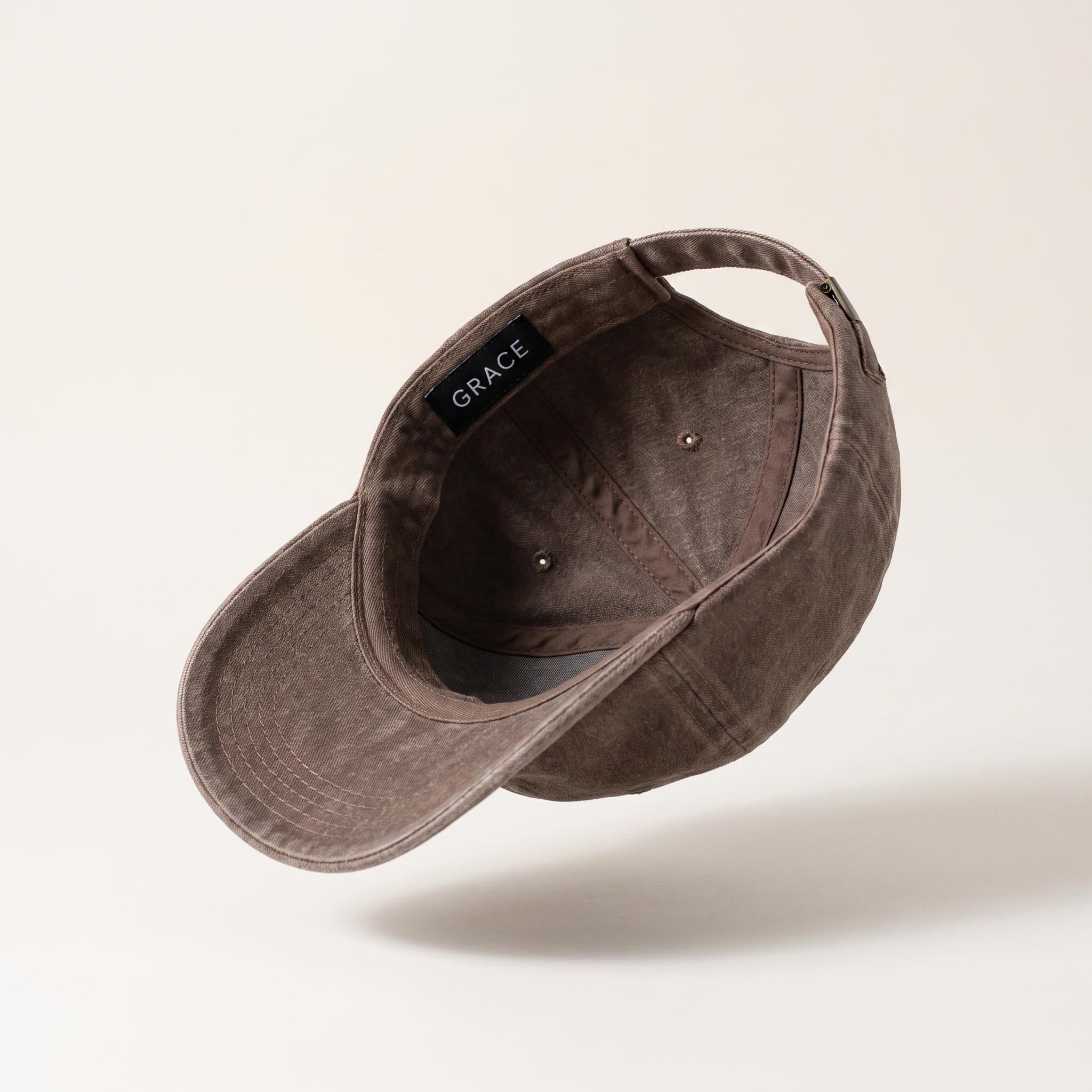 A washed dark brown baseball cap shown from an angled top-down perspective, floating against a light beige background with a soft shadow underneath. The brand name 'GRACE' is clearly visible on the black interior label.