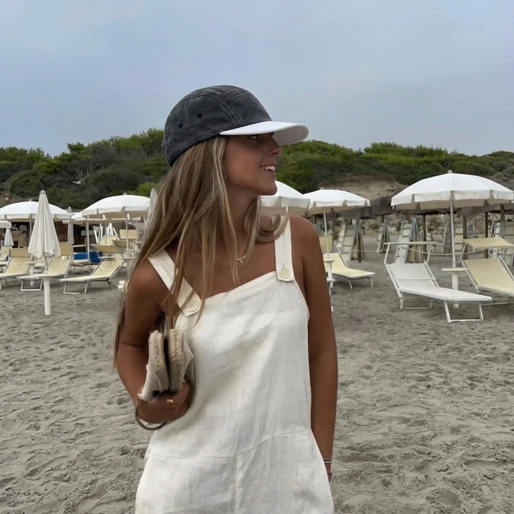 A woman standing on a sandy beach wearing a white linen one-shoulder dress, a dark gray baseball cap with a white brim, and holding a small beige clutch.