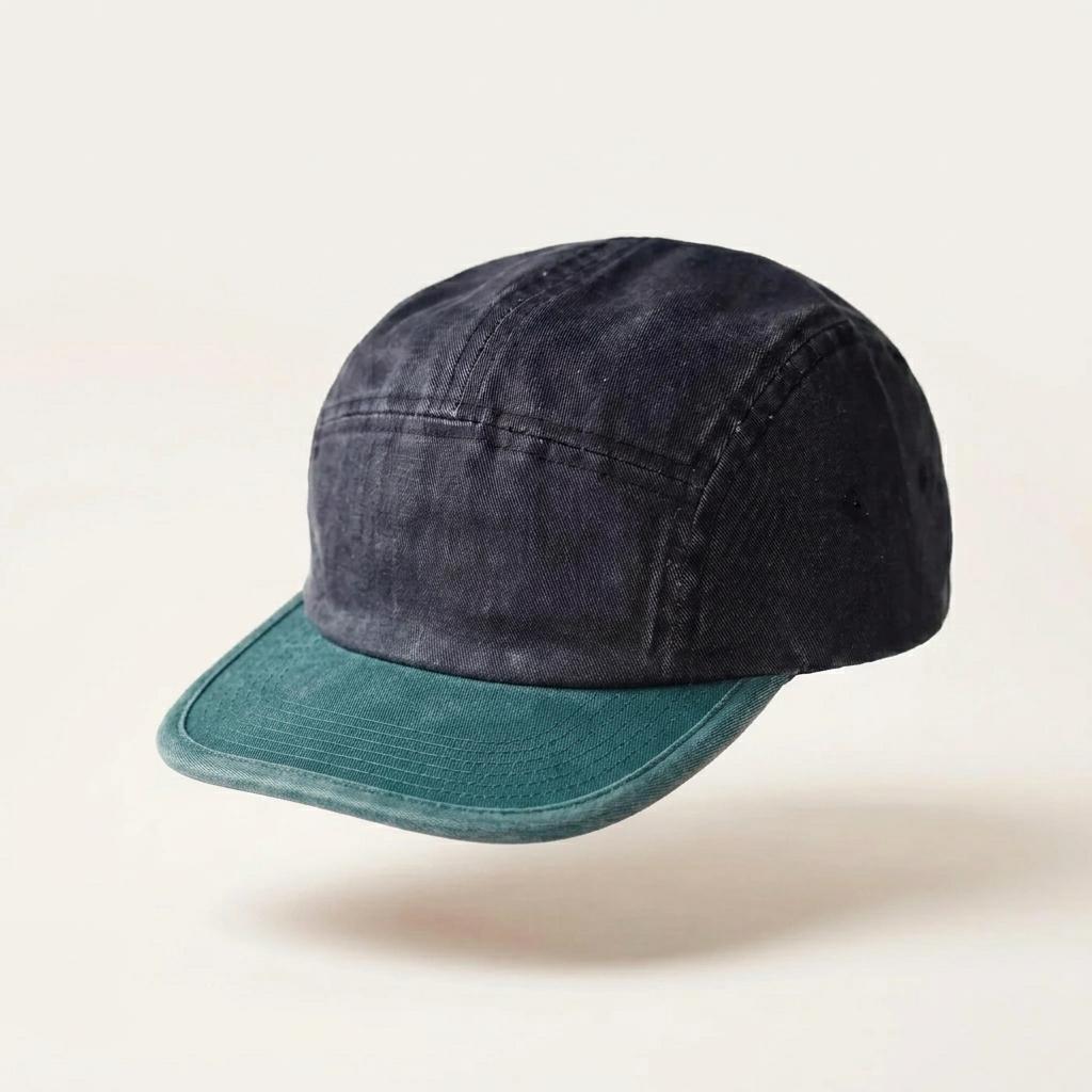 A five-panel camp cap with a dark navy blue washed cotton crown and a muted teal-green visor, shown floating against a light cream background.