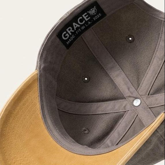 A structured baseball cap shown floating at an angle, featuring a dark gray fabric crown with a black interior label reading 'GRACE' and a contrasting tan suede-like visor.