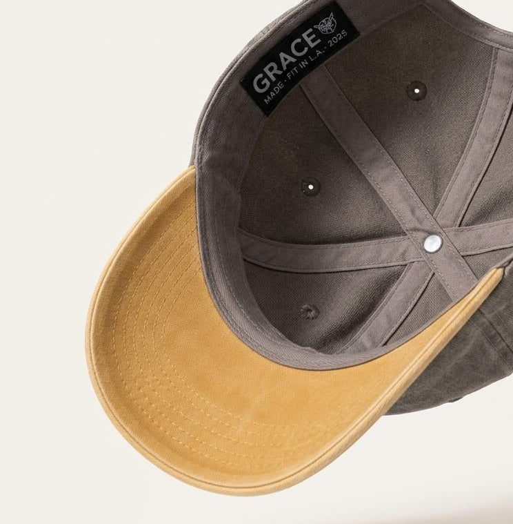 A structured baseball cap shown floating at an angle, featuring a dark gray fabric crown with a black interior label reading 'GRACE' and a contrasting tan suede-like visor.