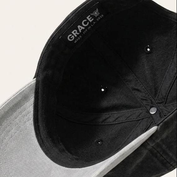 A black washed cotton baseball cap with a light gray curved brim, shown floating against a warm off-white background. The brand name 'Grace' is printed on the inside of the crown.