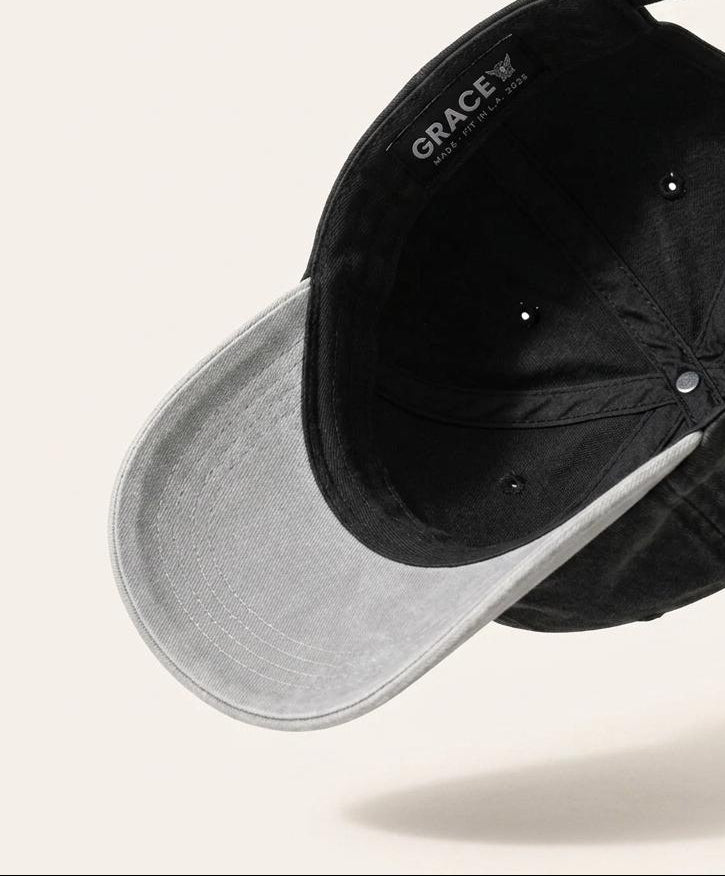 A black washed cotton baseball cap with a light gray curved brim, shown floating against a warm off-white background. The brand name 'Grace' is printed on the inside of the crown.