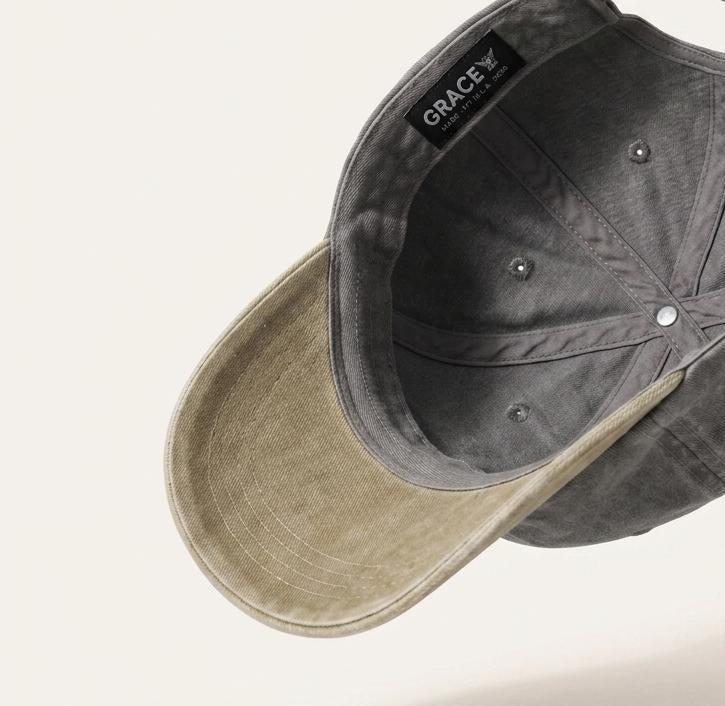 A dark gray washed cotton baseball cap with a light beige visor, shown floating against a light beige background. The inside of the crown features a black brand label reading 'ORACE'.