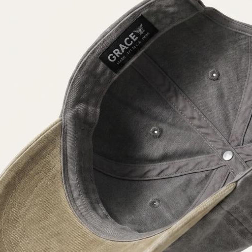A close-up angled shot of a baseball cap showing the dark gray fabric crown and the light beige brim, with a black interior label reading GRACE by Grace.