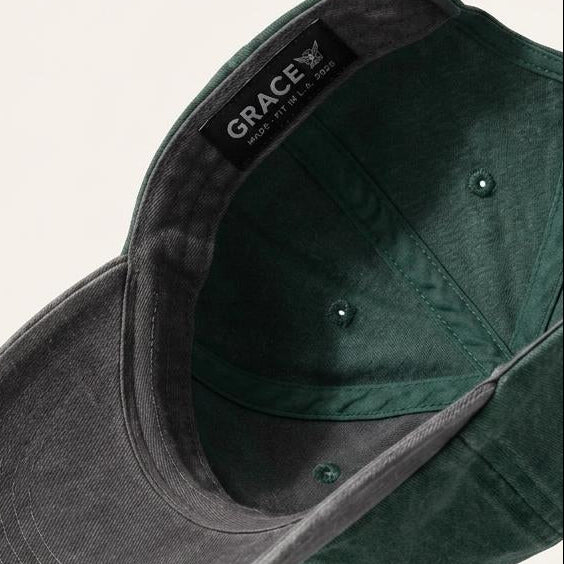 A dark forest green baseball cap with a washed, textured finish, shown floating at an angle to display both the curved brim and the interior. The brand name 'GRACE' is visible on a black label sewn inside the crown.