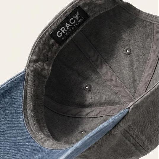 A dark gray washed cotton cap with a contrasting blue denim visor, shown floating against a light beige background. The brand name 'GRACK' is visible on the inner black label.