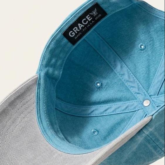 A light blue denim baseball cap shown from an angled top-down perspective, floating against a solid off-white background. The brand name 'GRACE' is clearly visible on a black label sewn inside the crown.