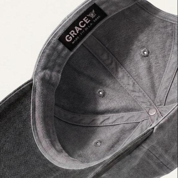 A dark gray washed cotton baseball cap shown floating at an angle, displaying the interior crown, snap closure, and a black interior label that reads 'GRACE'.