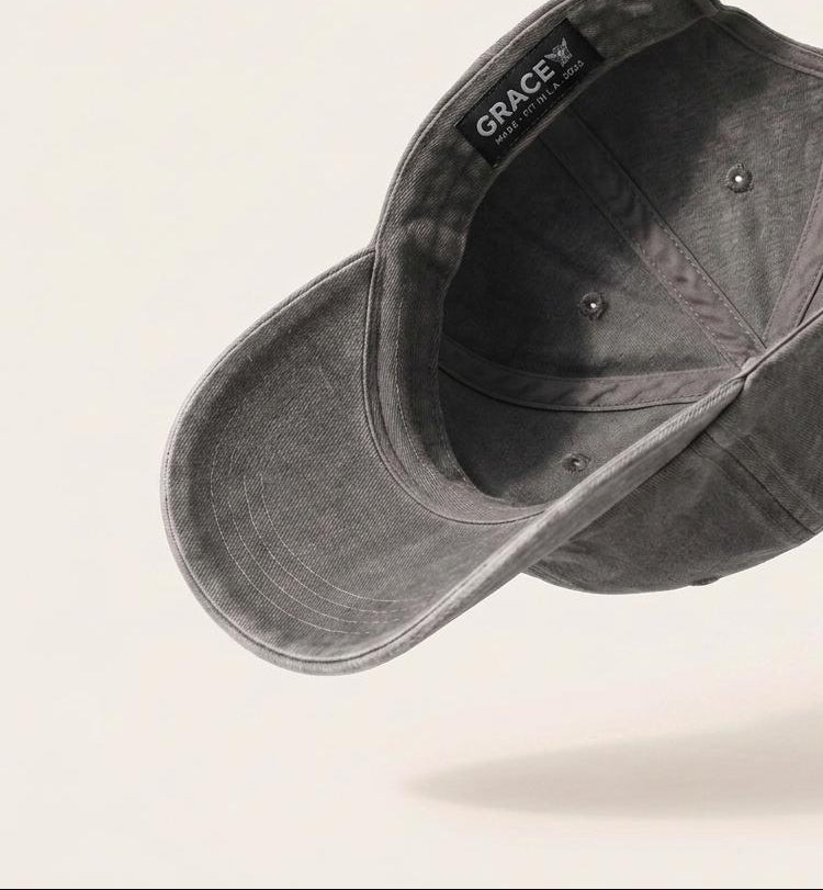 A dark gray washed cotton baseball cap floating at an angle against a light beige background, showing the interior with a black 'GRACE' brand label.