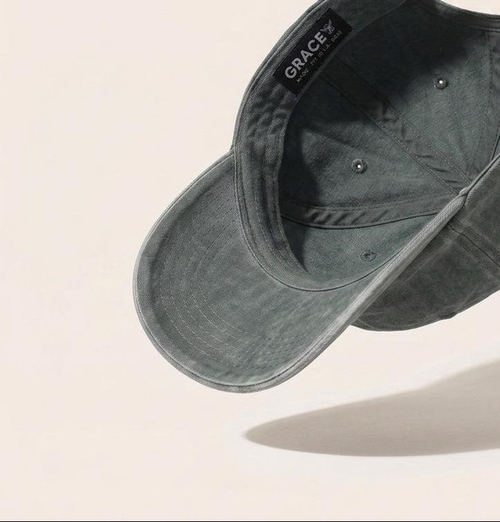 A dark green washed cotton baseball cap floating at an angle against a warm off-white background, with a black interior label reading 'GRACE'.