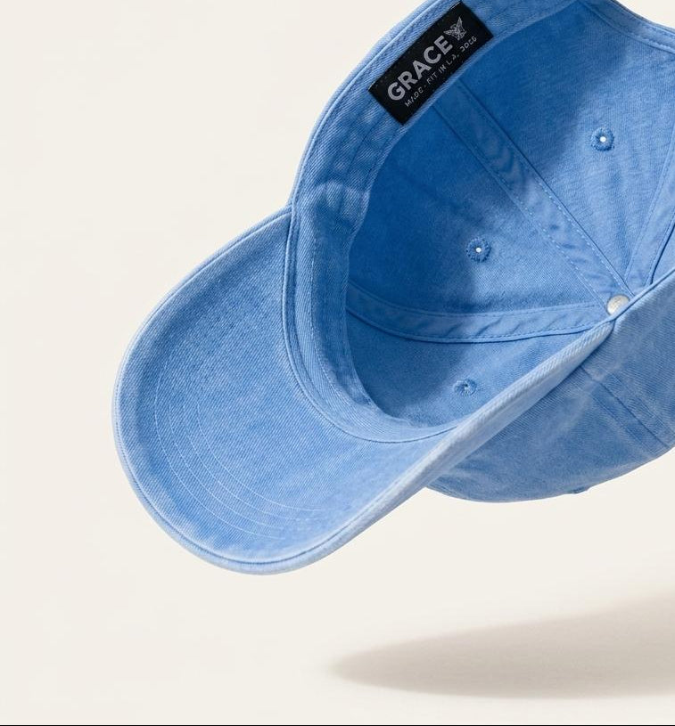 A light blue denim baseball cap shown from an angled top-down perspective, floating against a warm off-white background. The brand name 'GRACE' is clearly visible on a black label sewn onto the side of the cap.