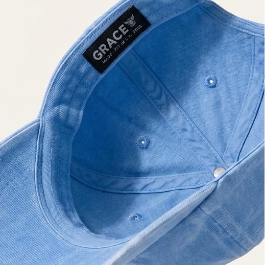A light blue denim baseball cap shown from an angled top-down perspective, floating against a warm off-white background. The brand name 'GRACE' is clearly visible on a black label sewn onto the side of the cap.