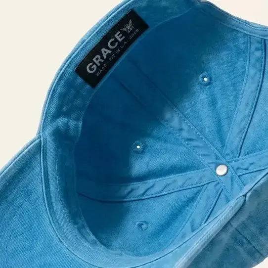 A light blue denim baseball cap shown from an angled top-down perspective, floating against a solid cream background. The brand name 'GRACE' is visible on a black interior label.